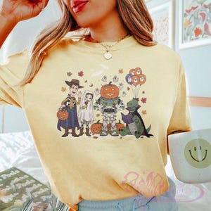 K&ouml;nnte beinhalten: Hellgelbes T-Shirt mit einem Halloween-Motiv mit Cartoon-Charakteren in Kost&uuml;men. Das Design zeigt Woody, ein Gespenst, Buzz Lightyear als K&uuml;rbis, einen Dinosaurier, der ein Buch liest, und K&uuml;rbisballons. Herbstbl&auml;tter und Geister sind ebenfalls enthalten.