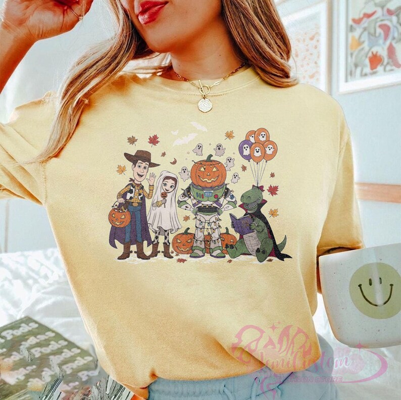 K&ouml;nnte beinhalten: Hellgelbes T-Shirt mit einem Halloween-Motiv mit Cartoon-Charakteren in Kost&uuml;men. Das Design zeigt Woody, ein Gespenst, Buzz Lightyear als K&uuml;rbis, einen Dinosaurier, der ein Buch liest, und K&uuml;rbisballons. Herbstbl&auml;tter und Geister sind ebenfalls enthalten.