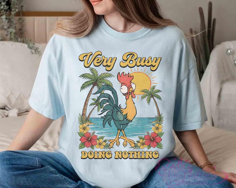 May include: Pale blue t-shirt featuring a cartoon rooster on a beach scene with palm trees and a sun. The text reads "Very Busy Doing Nothing". The shirt has a loose, comfortable fit.