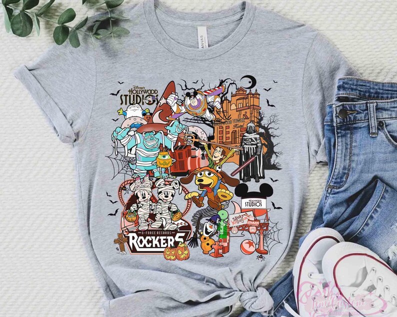 May include: A heather gray short-sleeve t-shirt featuring a colorful graphic print of various Disney characters in Halloween costumes, including Mickey Mouse, Buzz Lightyear, and Sulley, with the text Disney Hollywood Studios and Rockers.