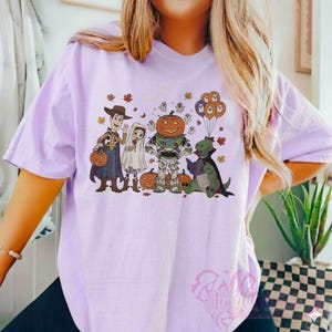 K&ouml;nnte beinhalten: Ein helllila T-Shirt mit einem Halloween-Motiv. Das Design zeigt Cartoon-Figuren in Kost&uuml;men: Woody, ein Geist, Buzz Lightyear mit einem K&uuml;rbiskopf und ein Dinosaurier, der ein Buch liest. Das Design enth&auml;lt auch Herbstbl&auml;tter und Geisterballons.