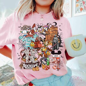 May include: A light pink t-shirt featuring a colorful graphic print of various Disney characters in Halloween costumes, including Mickey and Minnie Mouse, Sulley, and Woody. The design includes the text Disney Hollywood Studios and Rockers.