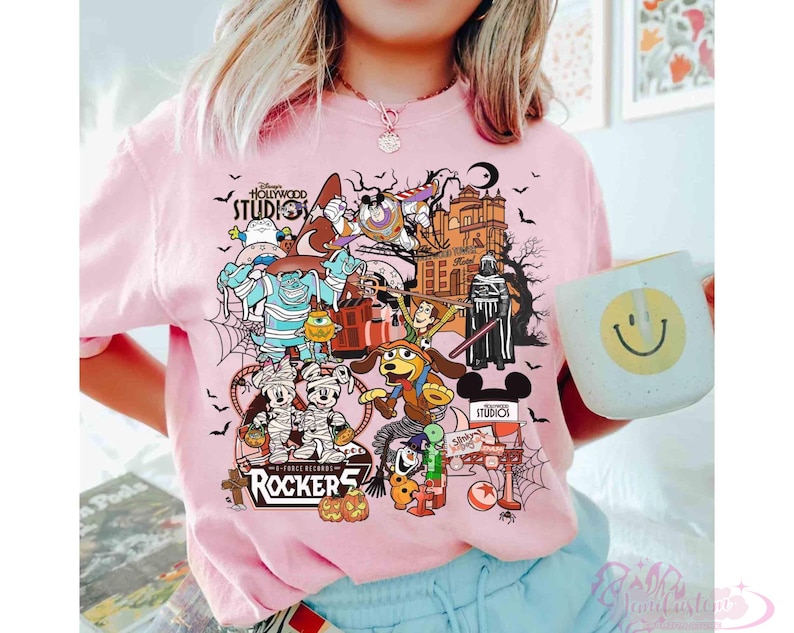 May include: A light pink t-shirt featuring a colorful graphic print of various Disney characters in Halloween costumes, including Mickey and Minnie Mouse, Sulley, and Woody. The design includes the text Disney Hollywood Studios and Rockers.