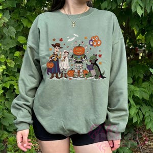 K&ouml;nnte beinhalten: Salbeigr&uuml;nes Sweatshirt mit einem Halloween-Motiv. Das Design zeigt Zeichentrickfiguren in Kost&uuml;men, darunter Woody, ein Geist, ein K&uuml;rbis und ein Dinosaurier, der ein Buch liest. Die Grafik enth&auml;lt auch Flederm&auml;use, Bl&auml;tter und Luftballons.