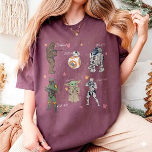 May include: A maroon t-shirt with illustrations of Star Wars characters: Chewbacca, BB-8, R2-D2, Boba Fett, Grogu, and a Stormtrooper. The characters' names are written above or below them in a whimsical font.