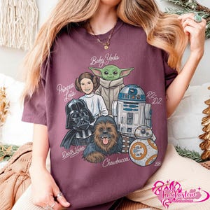 May include: Burgundy t-shirt featuring Star Wars characters: Baby Yoda, Princess Leia, Darth Vader, Chewbacca, R2-D2, and BB-8. The names of the characters are written above them in a stylized font. The shirt is a comfortable, casual wear item.