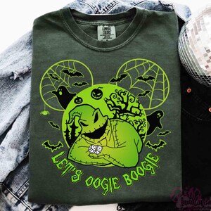 May include: Dark green t-shirt with a neon green graphic of a character with dice, bats, and spiderwebs. The text "Let's Oogie Boogie" is printed in neon green. The shirt is folded on top of a denim jacket and black trousers.