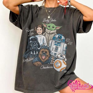 May include: Dark gray t-shirt featuring a graphic of Star Wars characters including Darth Vader, Princess Leia, Baby Yoda, Chewbacca, R2-D2, and BB-8. The names of the characters are printed above or below their images. The shirt has a relaxed fit.