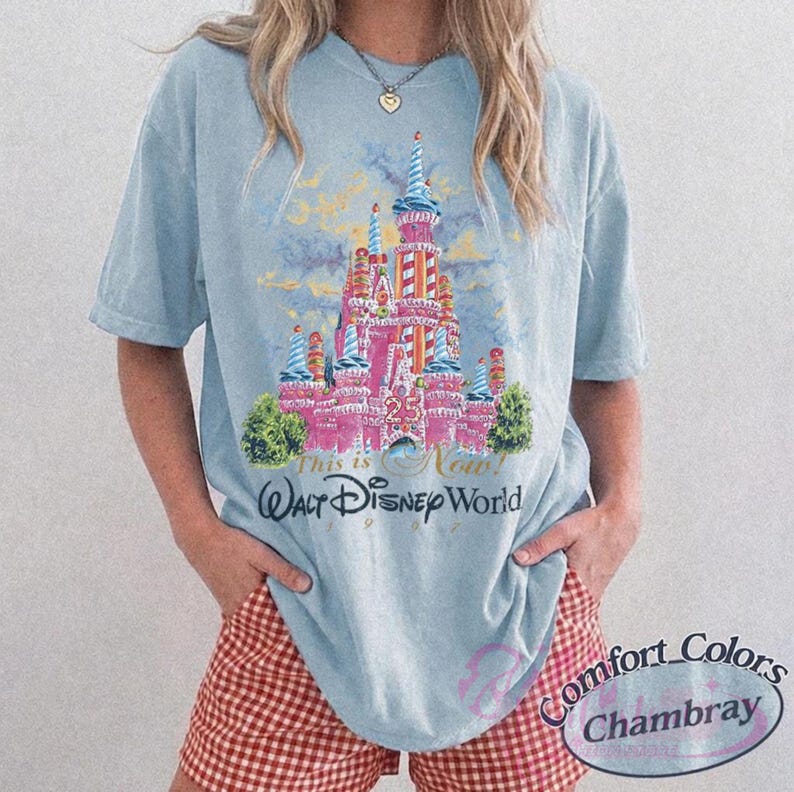 Op de afbeelding: Lichtblauw T-shirt met een roze kasteelafbeelding en de tekst "This is Now! Walt Disney World 1997". Het shirt heeft korte mouwen en een losse pasvorm. De afbeelding bevat het nummer "25".