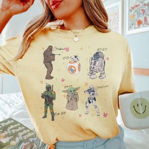 May include: Yellow t-shirt with illustrations of Star Wars characters: Chewbacca, BB-8, R2-D2, Boba Fett, Grogu, and a Stormtrooper. The design includes pink Mickey Mouse icons and character names.