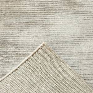 Op de afbeelding: Een close-up van een beige tapijt met een getextureerd, horizontaal gestreept patroon. De hoek van het tapijt is opgetild en onthult de geweven achterkant. Het tapijt heeft een neutraal kleurenpalet, geschikt voor diverse interieurstijlen.