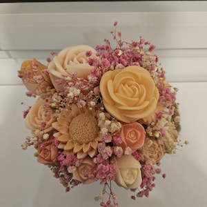 Peut inclure: Un bouquet de fleurs en bois sculpté aux tons pêche, crème et marron clair, agrémenté de gypsophile rose et blanc séché. L'arrangement comprend des roses et un tournesol.