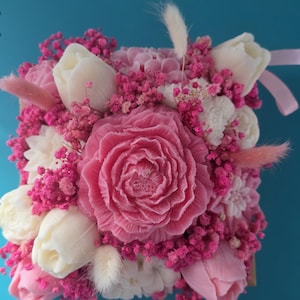 Peut inclure: Un arrangement décoratif carré composé d'une grande bougie centrale en forme de rose rose, entourée de bougies en forme de tulipes blanches, de petites fleurs séchées roses et de plumes d'herbe duveteuses rose clair dans un contenant carré.