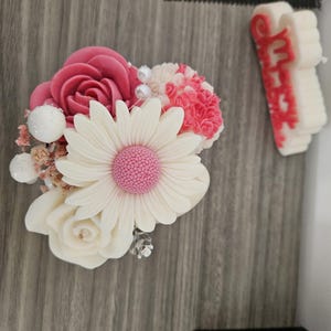 Peut inclure: Composition florale décorative avec une grande marguerite blanche au cœur rose, entourée de roses et d'œillets roses et blancs. L'arrangement comprend de petites boules décoratives blanches et des perles, créant une esthétique délicate.