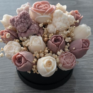 Könnte beinhalten: Ein Blumenarrangement aus Seifenrosen in Rosa-, Mauve- und Weißtönen, präsentiert in einer runden schwarzen Box. Das Bouquet umfasst verschiedene Rosenstile und wird durch kleine, getrocknete Blumen ergänzt.