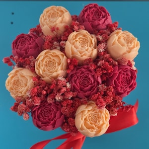 Può includere: Un bouquet di saponi modellati a forma di fiore nelle tonalità pesca e magenta intenso, intervallati da piccoli grappoli di gipsofila rossa essiccata. La composizione è legata alla base con un nastro di raso rosso brillante.