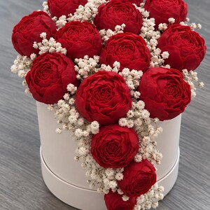 Puede incluir: Una caja de regalo cilíndrica blanca llena de un arreglo denso de flores de color rojo intenso similares a las peonías y pequeñas flores blancas de paniculata. El arreglo floral está agrupado en la parte superior y cae en cascada.