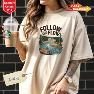 May include: Ivory-colored t-shirt with the text "FOLLOW THE FLOW" above a graphic of a river winding through a forest. The shirt is a Comfort Colors 1717 style. A clear plastic cup with a green logo is held in the image.