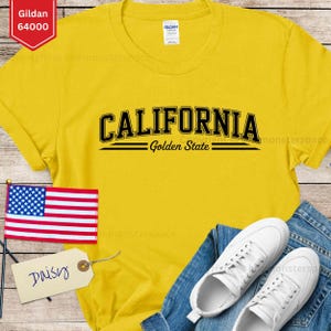 May include: A yellow t-shirt with the word "CALIFORNIA" in black arched lettering, with "Golden State" below. An American flag and a tag that says "Daisy" are also visible. The shirt is paired with blue jeans and white sneakers.