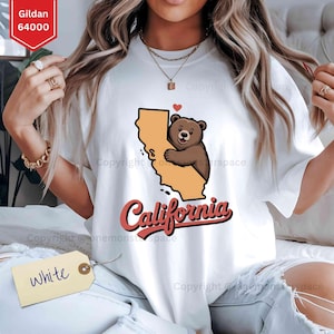 May include: White t-shirt featuring a graphic of a brown bear hugging the state of California, with a red heart above the bear. The word "California" is written in a cursive font below the graphic. The shirt has a tag that says "White".
