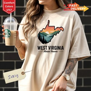 May include: Ivory-colored t-shirt featuring a West Virginia design. The graphic includes the state outline with a sunset, mountains, and trees. The text "WEST VIRGINIA Almost Heaven" is printed below the graphic. The shirt has a relaxed fit.