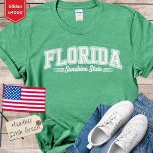 May include: A green t-shirt with the word "FLORIDA" in white block letters and "Sunshine State" underneath. The shirt is paired with blue jeans and white trainers. An American flag and a tag that says "Heather Irish Green" are also in the image.