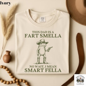 May include: Ivory-colored t-shirt with the text "THIS DAD IS A FART SMELLA, NO WAIT, I MEAN SMART FELLA." A cartoon frog wearing a cowboy hat and boots, holding a gun, is printed on the front. The shirt is made of Comfort Colors material.