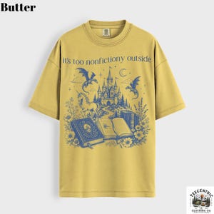May include: Butter-colored t-shirt with a blue graphic featuring a castle, dragons, a unicorn, and open books. The text "it's too nonfictiony outside" is printed above the graphic. The shirt has a crew neck and short sleeves.