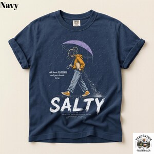 May include: A navy blue t-shirt featuring a graphic of a person in a yellow hoodie walking under a purple umbrella in the rain. The text reads All these FLAVORS and you choose to be SALTY, along with smaller text below the word SALTY.