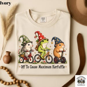 May include: An ivory t-shirt featuring a graphic of an opossum on a tricycle, a frog on a bicycle, and a hamster on a hoverboard, all wearing pointed hats. The text below reads, Off To Cause Maximum Kerfuffle, surrounded by small stars.