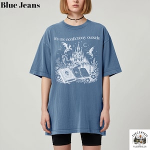 May include: Blue short-sleeved t-shirt with the text "it's too nonfictiony outside" above a white graphic of a castle, dragons, and books. The shirt is a faded blue color.