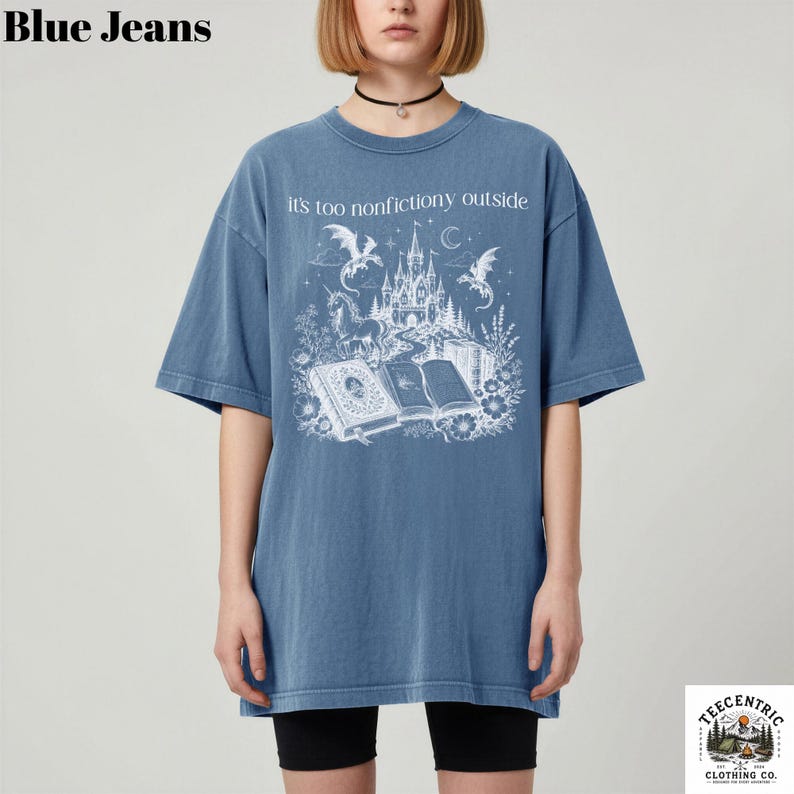 May include: Blue short-sleeved t-shirt with the text "it's too nonfictiony outside" above a white graphic of a castle, dragons, and books. The shirt is a faded blue color.