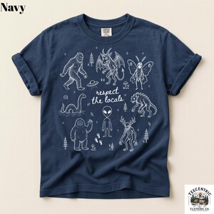 May include: A navy blue short-sleeve t-shirt featuring a white line-art graphic of various cryptids, including Bigfoot, a mothman, an alien, and the Loch Ness monster, with the text "respect the locals" printed in the center of the design.