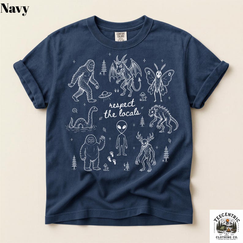 May include: A navy blue short-sleeve t-shirt featuring a white line-art graphic of various cryptids, including Bigfoot, a mothman, an alien, and the Loch Ness monster, with the text "respect the locals" printed in the center of the design.