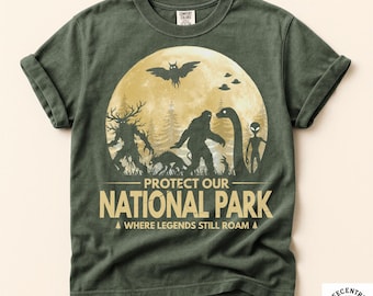 May include: A moss green t-shirt featuring a graphic of a large moon, a forest, and various cryptids including Bigfoot, an alien, and a mothman. The text reads PROTECT OUR NATIONAL PARK WHERE LEGENDS STILL ROAM. The shirt has a crew neck and short sleeves.