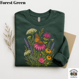 May include: A forest green crewneck sweatshirt featuring a colorful botanical illustration of wildflowers, a monarch butterfly, a dragonfly, and a bee. The graphic includes pink coneflowers, yellow daisies, and purple lavender stalks.