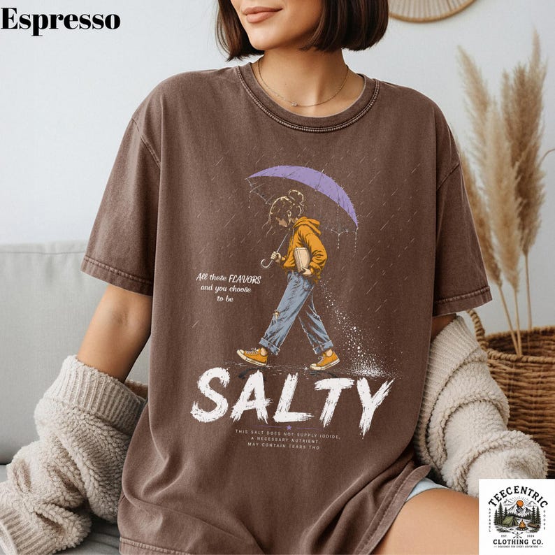 May include: A brown short-sleeve t-shirt featuring a graphic of a person holding a purple umbrella while walking in the rain. The text reads All these FLAVORS and you choose to be SALTY, along with smaller text below. The shirt has a crew neckline.