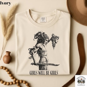 May include: Ivory-colored crew neck t-shirt with a black and white illustration of a woman holding a sword and a severed head. The text "GIRLS WILL BE GIRLS" is printed below the illustration. The shirt is from Comfort Colors.