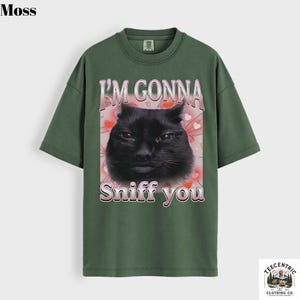 May include: A moss green t-shirt with a graphic of a black cat and the text "I'M GONNA Sniff you" in pink. The design includes pink hearts. The shirt has a crew neck and short sleeves. The brand logo is in the bottom right corner.