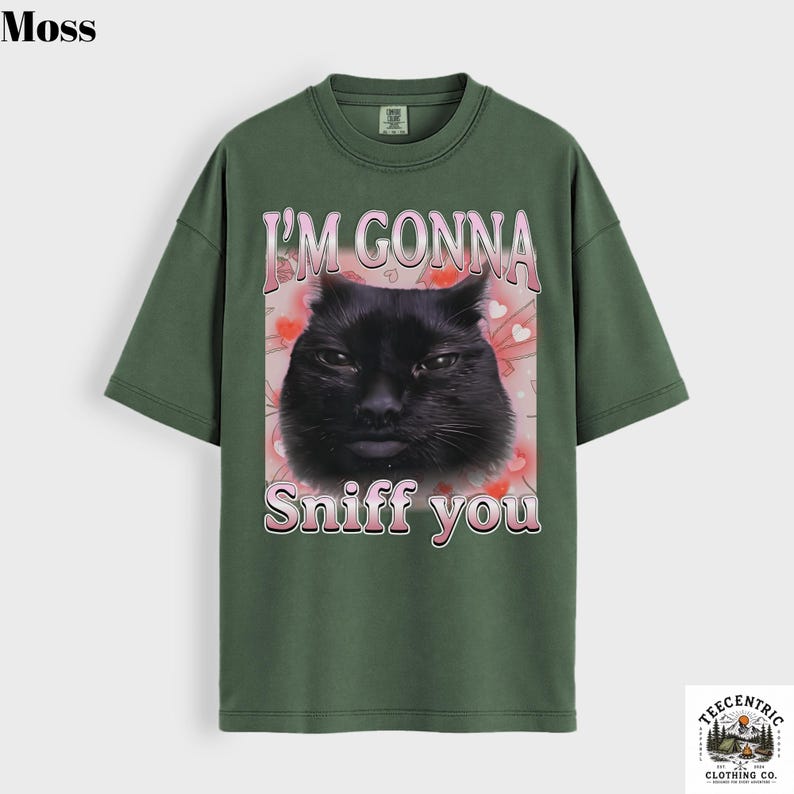 May include: A moss green t-shirt with a graphic of a black cat and the text "I'M GONNA Sniff you" in pink. The design includes pink hearts. The shirt has a crew neck and short sleeves. The brand logo is in the bottom right corner.