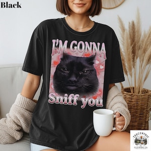 May include: Black t-shirt featuring the text "I'M GONNA Sniff you" and a graphic of a black cat's face. The cat graphic is set against a pink background with small hearts. The shirt is a casual style.