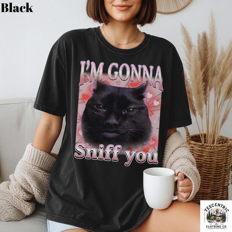 May include: Black t-shirt featuring the text "I'M GONNA Sniff you" and a graphic of a black cat's face. The cat graphic is set against a pink background with small hearts. The shirt is a casual style.