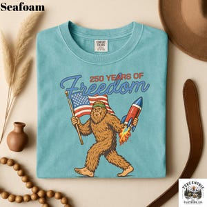 K&ouml;nnte beinhalten: T&uuml;rkisfarbenes T-Shirt mit einem Cartoon-Bigfoot, der eine amerikanische Flagge und eine Rakete h&auml;lt. Der Text "250 YEARS OF Freedom" befindet sich &uuml;ber dem Bigfoot. Das T-Shirt hat einen Rundhalsausschnitt und ein Comfort Colors-Etikett.