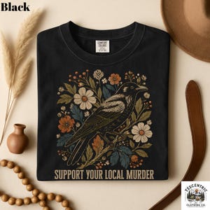 May include: Black t-shirt featuring a detailed illustration of a crow perched on a branch, surrounded by floral elements in shades of orange, white, and green. The text "SUPPORT YOUR LOCAL MURDER" is printed below the design. A Comfort Colors tag is visible.