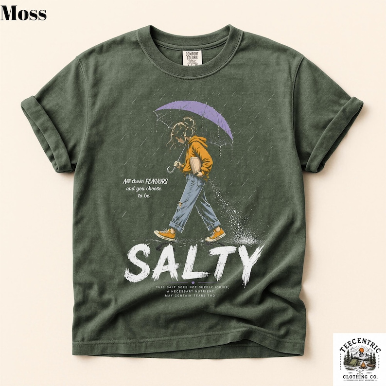 May include: A moss green t-shirt featuring a graphic of a person walking in the rain under a purple umbrella. The text reads All these FLAVORS and you choose to be SALTY, along with smaller text below. The shirt has a crew neck and short sleeves.