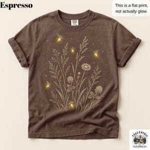 May include: An espresso-colored short-sleeve t-shirt featuring a graphic print of wildflowers and glowing fireflies. The design includes text stating Espresso and This is a flat print, not actually glow. The shirt has a crew neckline and rolled sleeves.