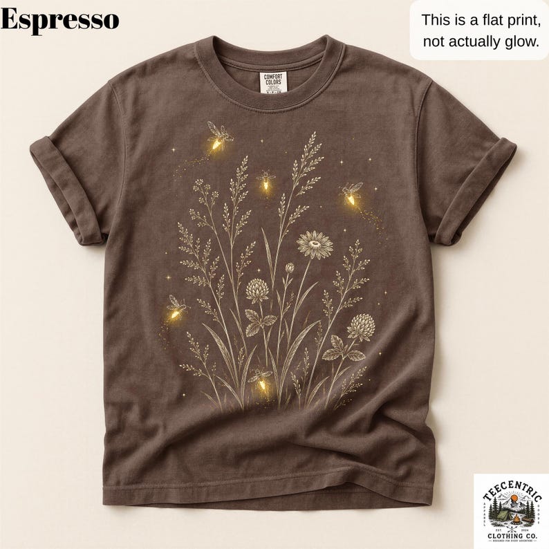 May include: An espresso-colored short-sleeve t-shirt featuring a graphic print of wildflowers and glowing fireflies. The design includes text stating Espresso and This is a flat print, not actually glow. The shirt has a crew neckline and rolled sleeves.