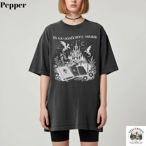 May include: Dark gray t-shirt with a fantasy graphic. The graphic features a castle, books, a unicorn, and dragons, with the text "it's too nonfictiony outside". The shirt is a relaxed fit and has short sleeves.