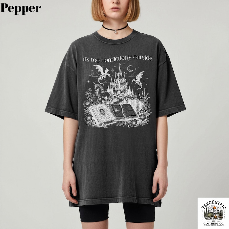 May include: Dark gray t-shirt with a fantasy graphic. The graphic features a castle, books, a unicorn, and dragons, with the text "it's too nonfictiony outside". The shirt is a relaxed fit and has short sleeves.