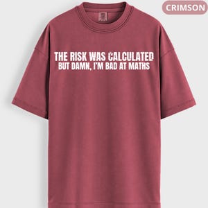 Peut inclure: T-shirt de couleur cramoisie avec l'inscription blanche : "THE RISK WAS CALCULATED BUT DAMN, I'M BAD AT MATHS". Le t-shirt a un col rond et des manches courtes. Le mot "CRIMSON" est dans un encadr&eacute; en haut &agrave; droite.
